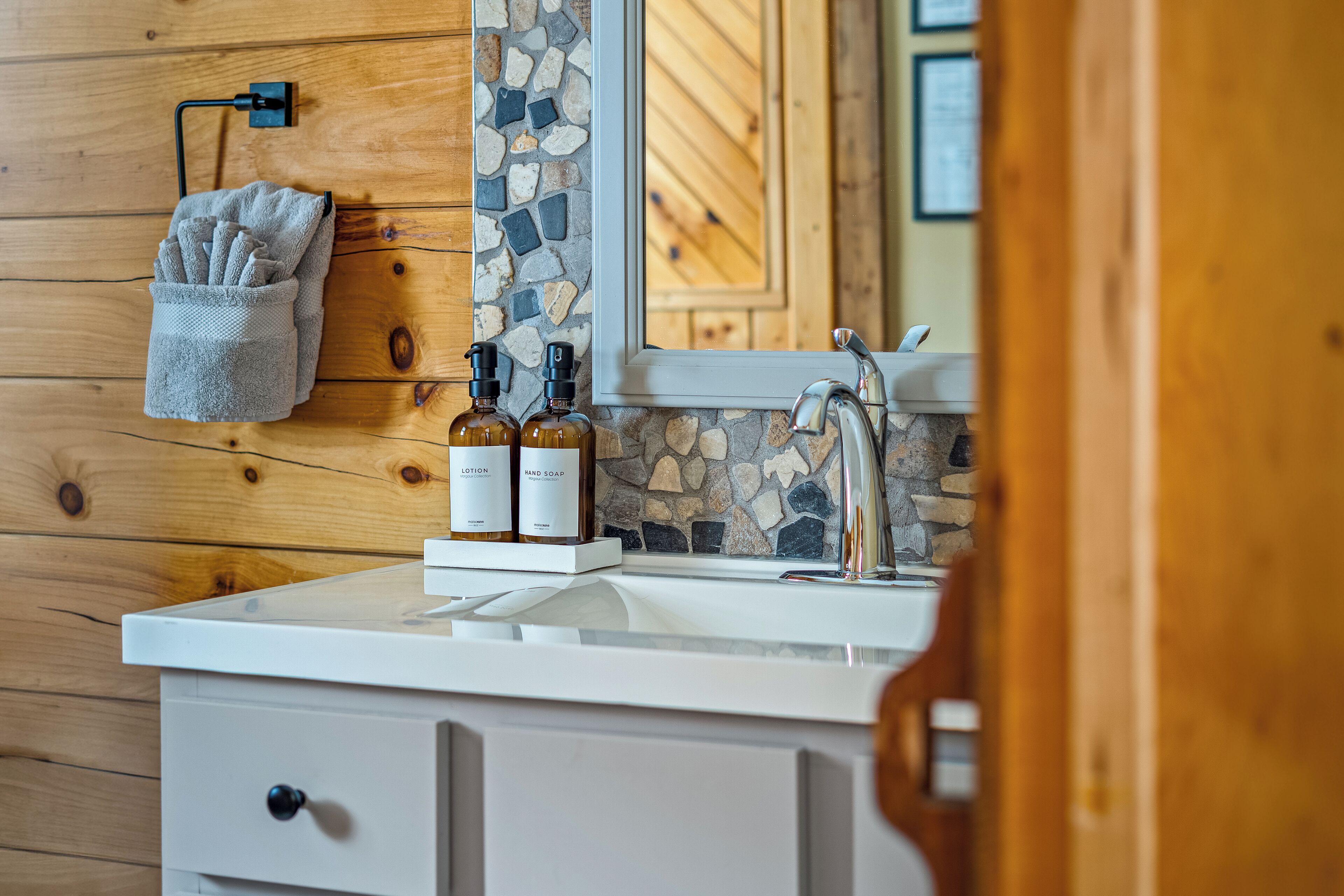 A bathroom with a stone mosaic backsplash, a wooden wall, a towel rack, and a sink with two bottles of soap.