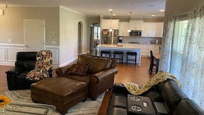 A modern living room with a brown leather sofa, a black leather chair, and a patterned throw. A kitchen with white cabinets and stainless steel appliances is visible in the background.