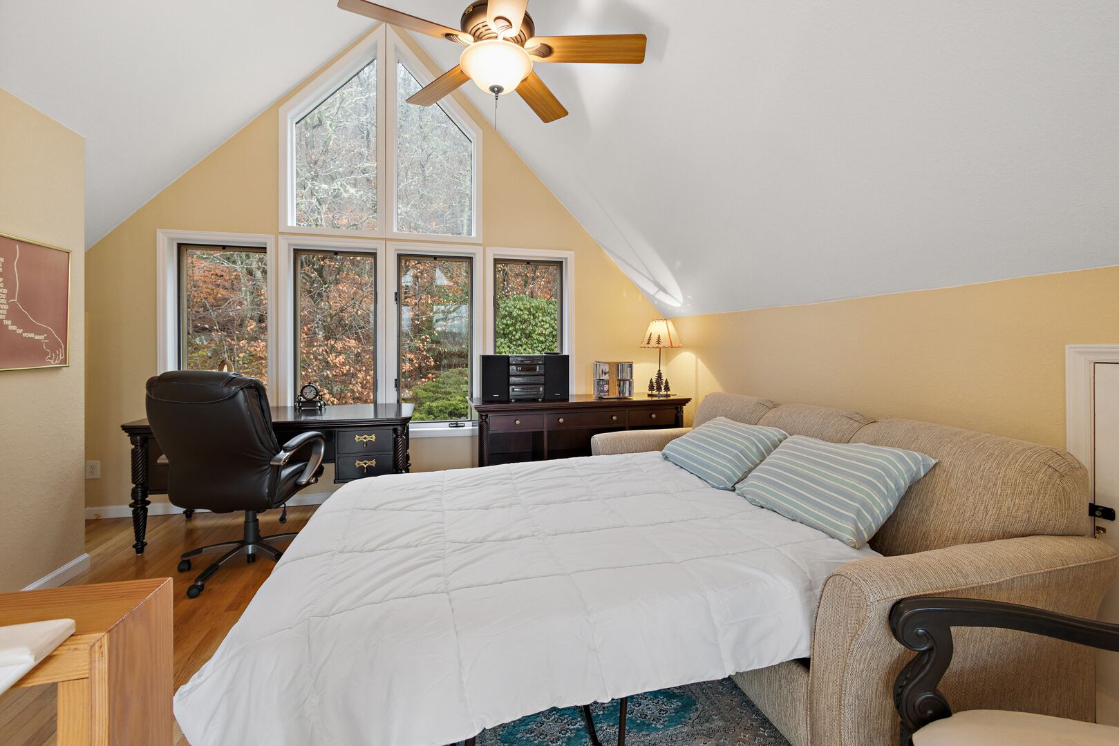 A bedroom with a large bed, a desk, and a chair. There are multiple windows providing a view of trees.
