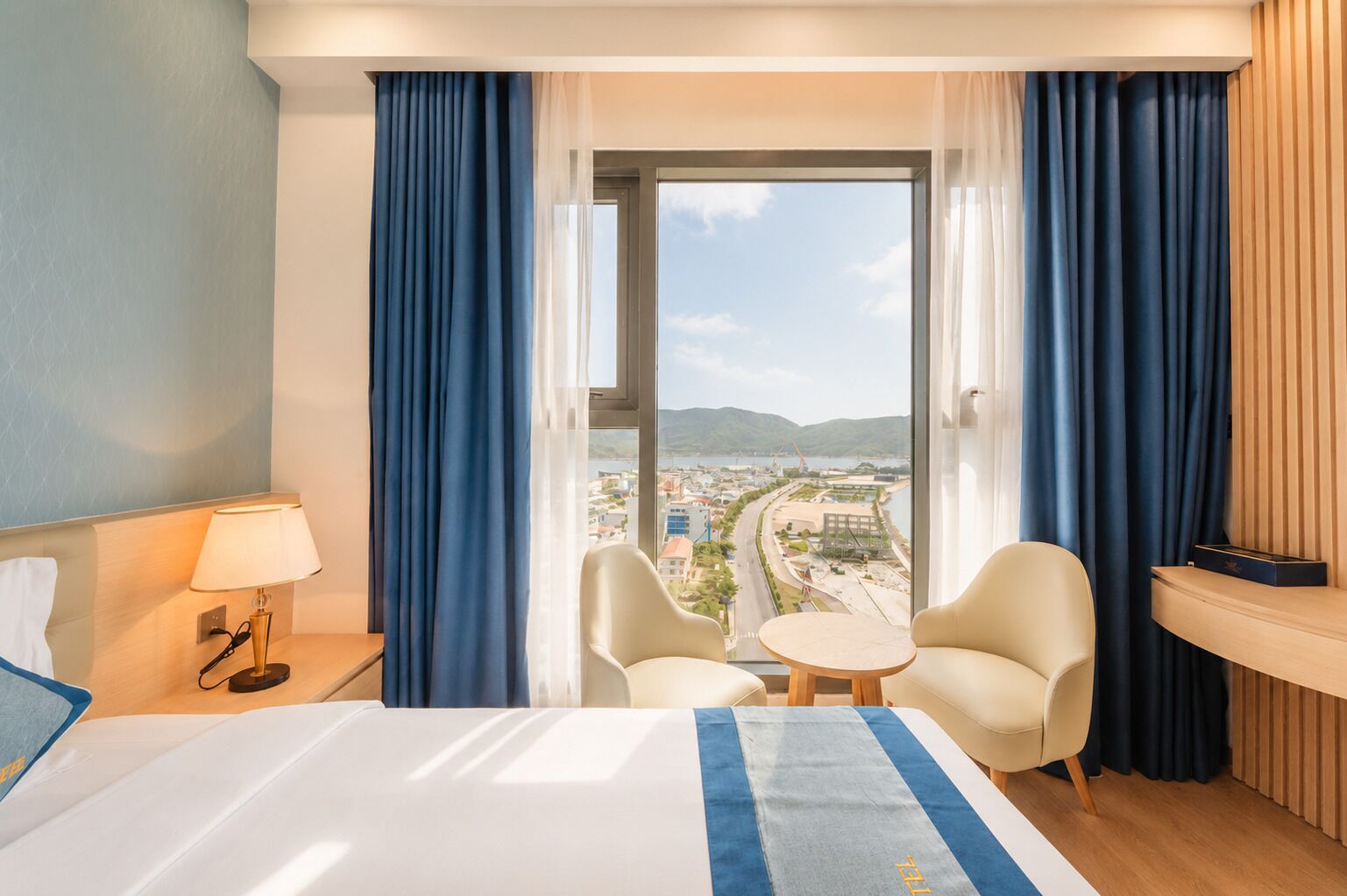 A hotel room with a bed, two chairs, a small table, a lamp, and a view of a cityscape through a large window.