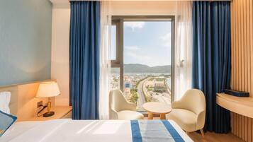 A hotel room with a bed, two chairs, a small table, a lamp, and a view of a cityscape through a large window.