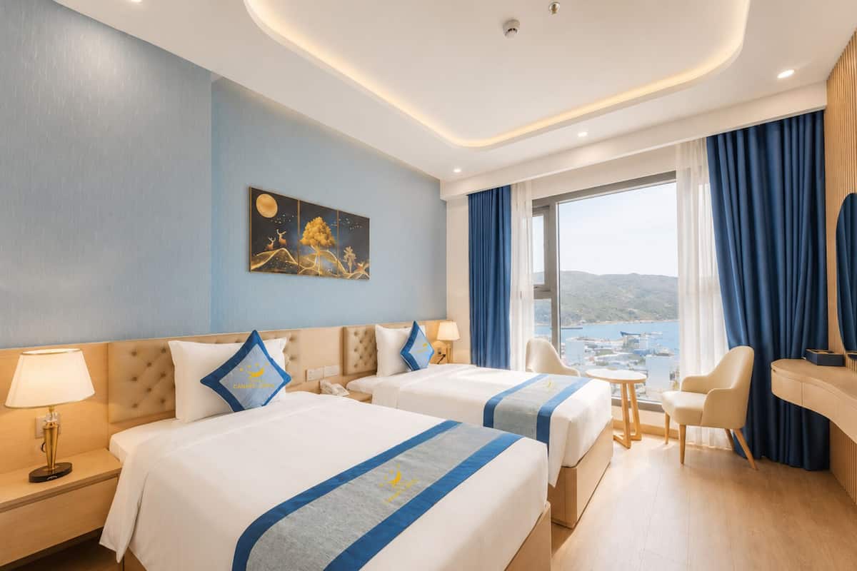 Habitación de hotel con dos camas, un escritorio, una silla, vista al mar y un cuadro en la pared.
