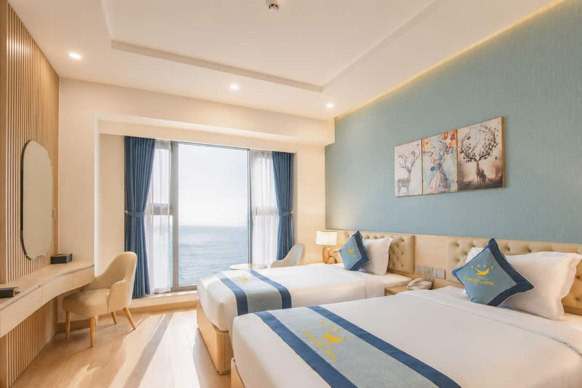 Habitación de hotel con dos camas, un escritorio, una silla, un ventanal con cortinas y obras de arte en la pared.