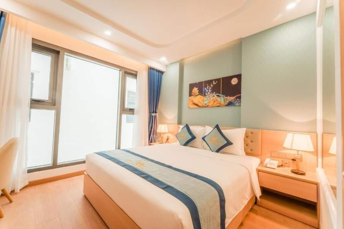 Una habitación de hotel con una cama grande, una mesita de noche con lámpara, una silla y una ventana con cortinas.