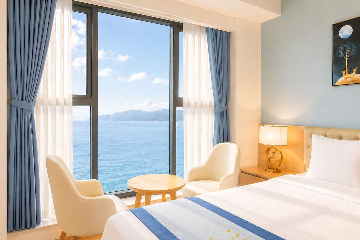 Habitación de hotel con cama, dos sillas, una mesita, una mesita de noche con lámpara y un ventanal con vista al mar.