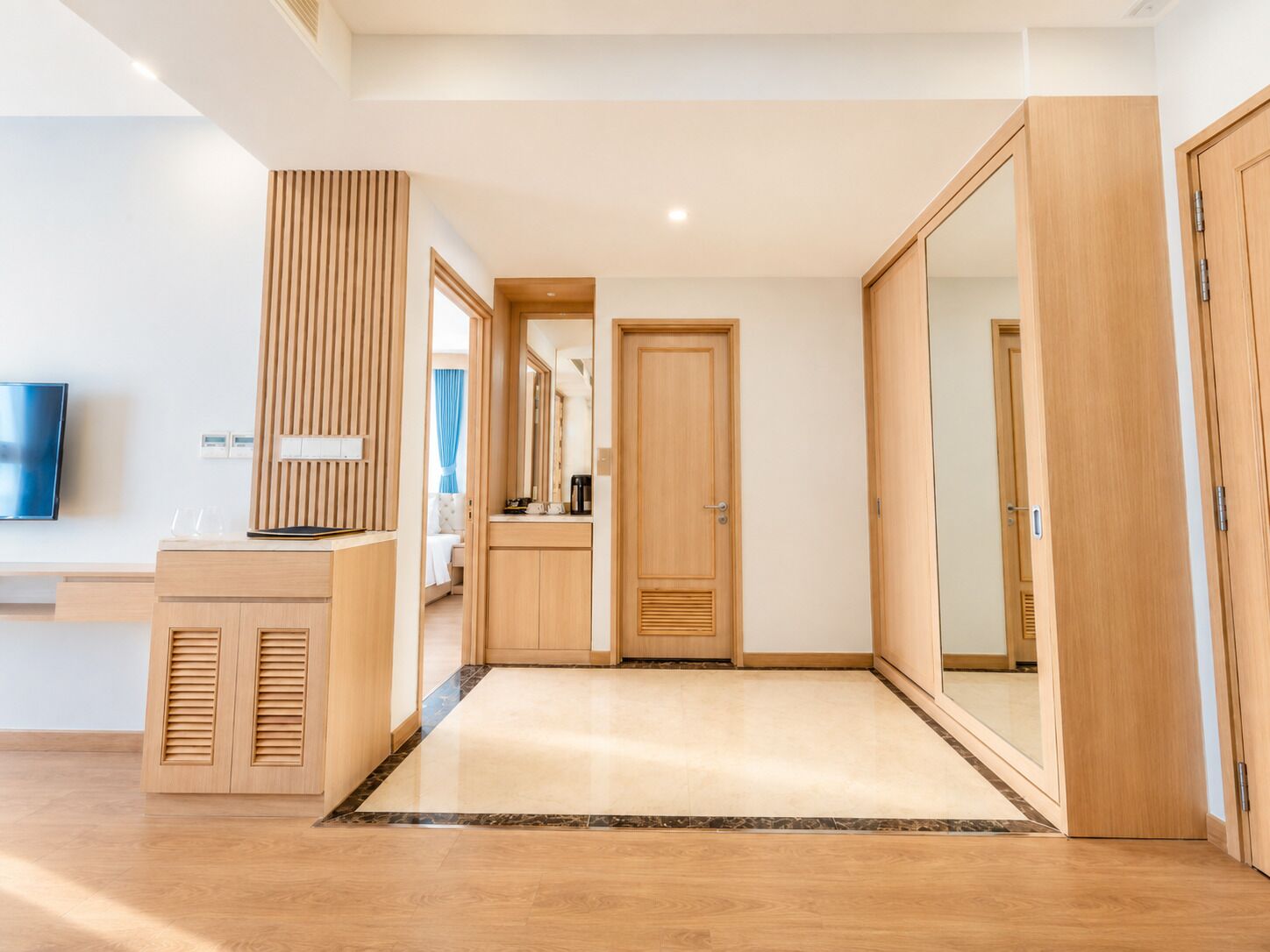 A modern hotel room with a wooden wardrobe, a desk with a flat-screen TV, and a large mirror.