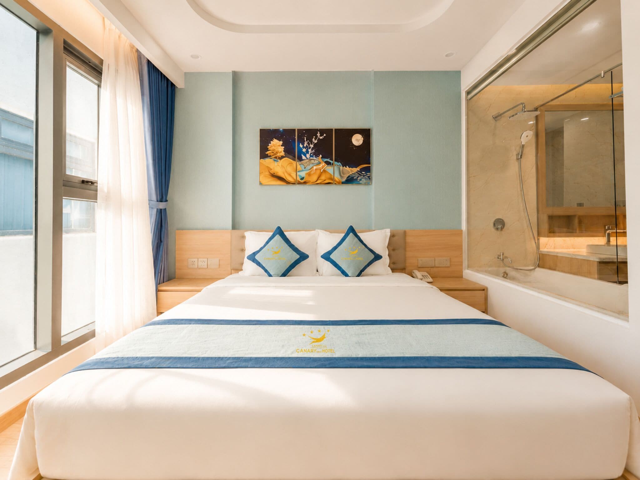 A hotel room with a bed, a nightstand, a painting on the wall, and a bathroom with a shower.