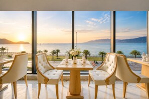 A dining area with a table and chairs, a vase with flowers, and a view of the sea and mountains. - Canary Gold Hotel Quy Nhơn, Beachfront (Quy Nhon)