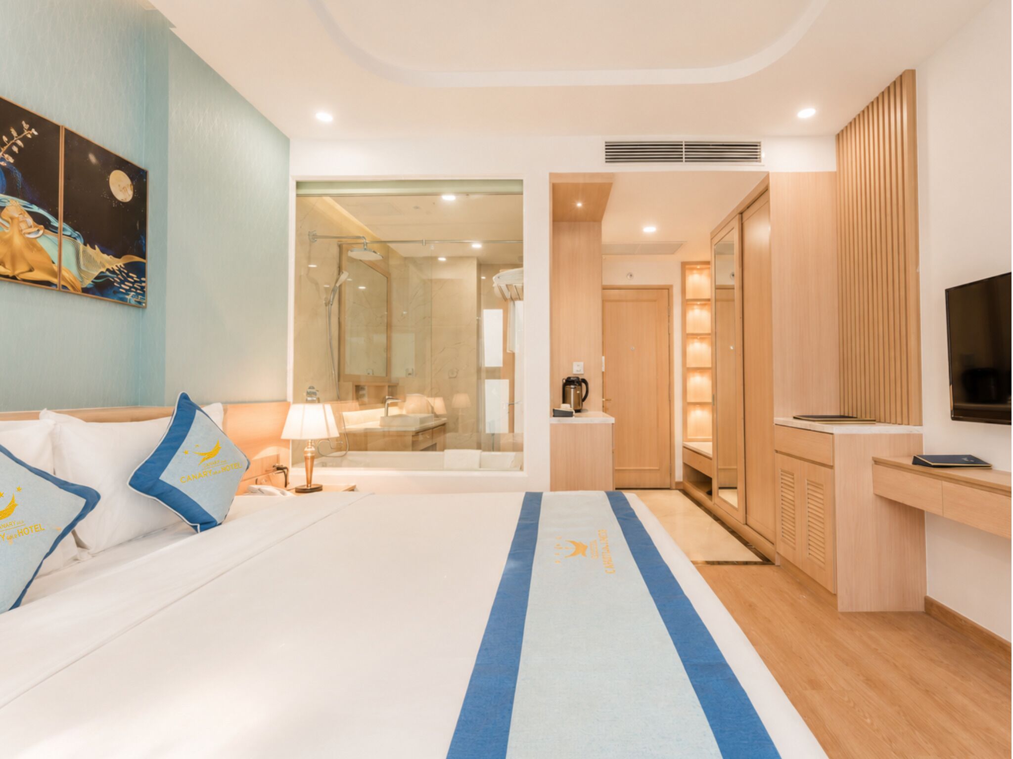A hotel room with a bed, a desk, a TV, a bathroom with a mirror, and a painting on the wall.