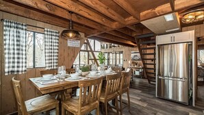 Ein Holztisch ist gedeckt mit Tellern, Gläsern und Besteck, umgeben von Holzstühlen. Im Hintergrund ist ein Edelstahl-Kühlschrank zu sehen.