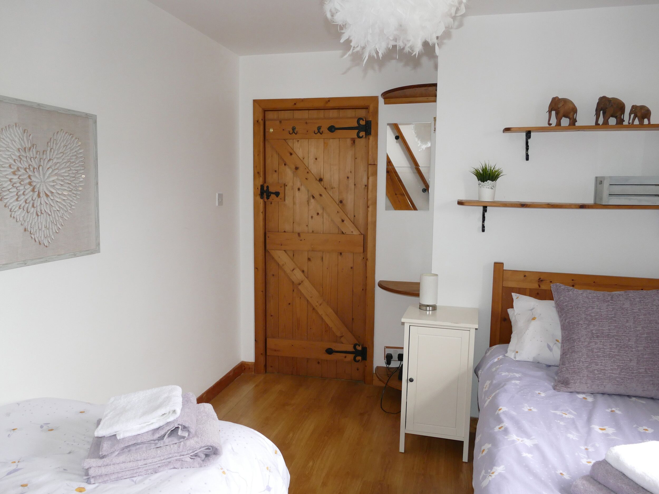 Un dormitorio con puerta de madera, cama con cubrecama floral, mesita de noche con lámpara y estante con adornos.