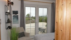 Un dormitorio con cama, una silla, mesita de noche, planta en maceta, estante de pared con objetos decorativos y un ventanal con vistas al exterior.