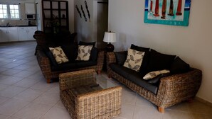 A living room with wicker furniture, a painting on the wall, and a kitchen in the background. - Villa Olivia Poolside, Dive villa, 300 m from the Ocean! (Kralendijk)