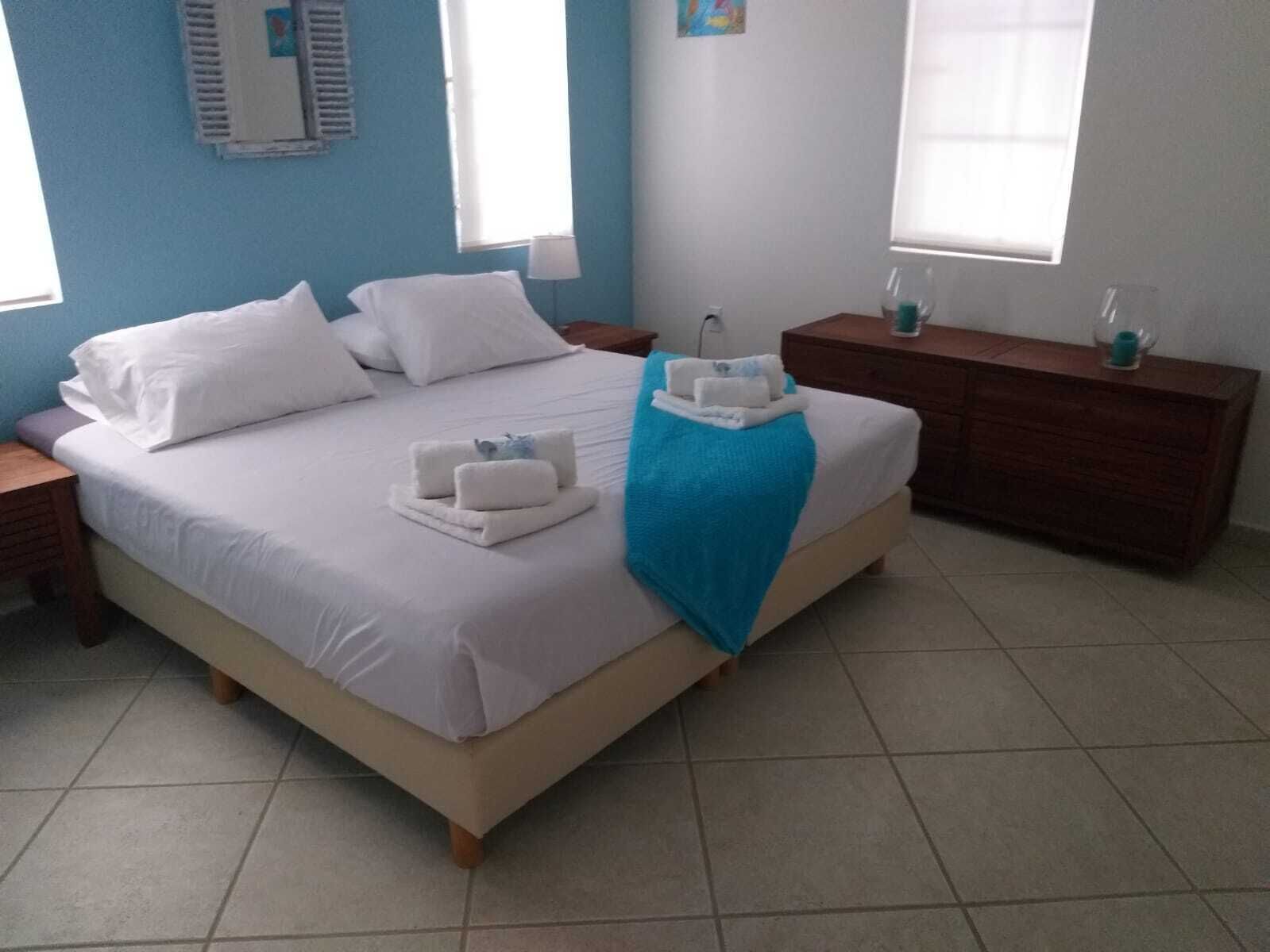 A neatly made bed with white linens and towels, a blue throw blanket, and a wooden nightstand with a lamp.