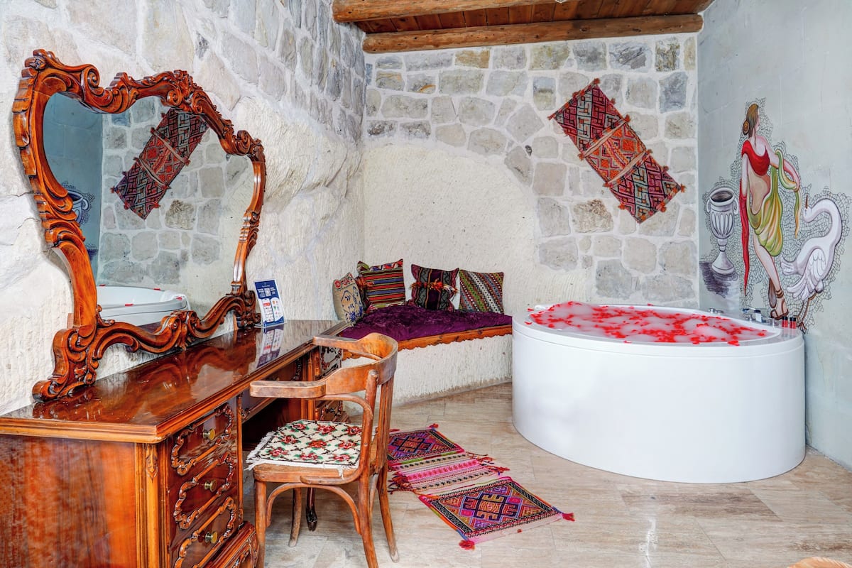 A room with a stone wall, a wooden vanity with a mirror, a white bathtub, a wooden chair, and a patterned rug.