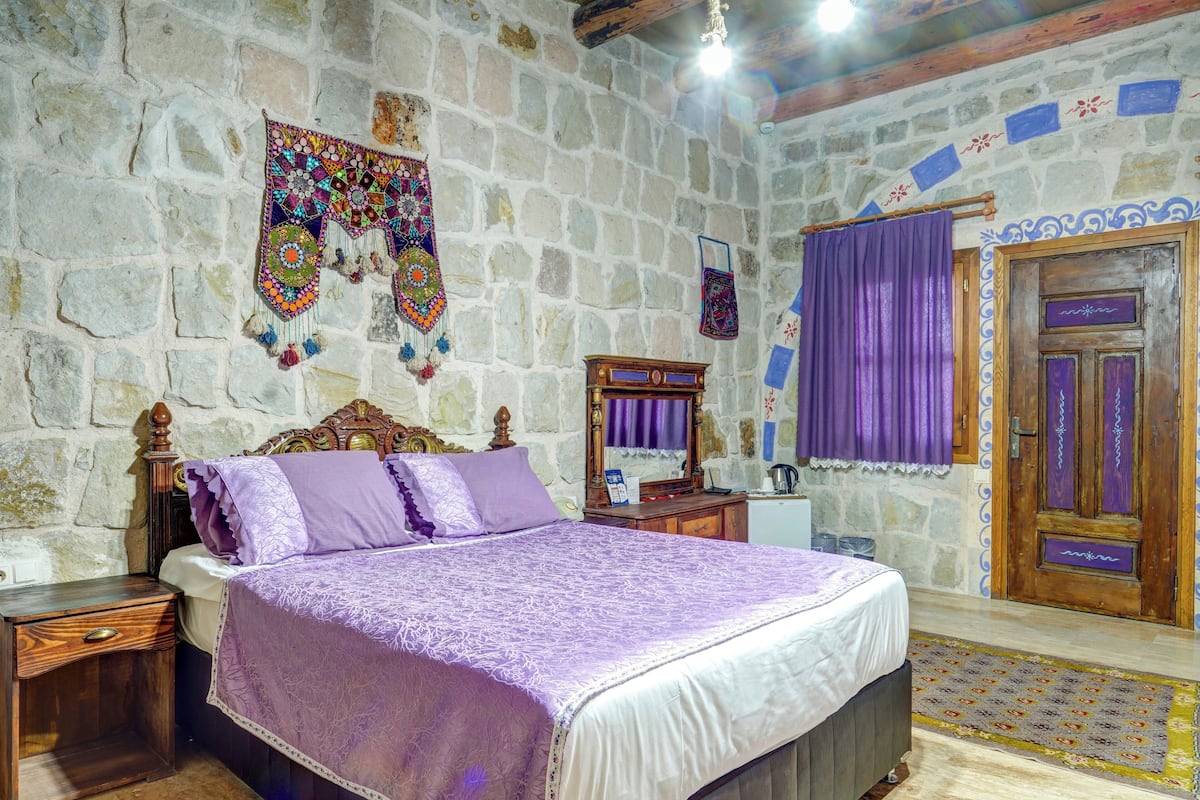 A room with a stone wall, a bed with a purple bedspread, a wooden nightstand, a wooden dresser, and a door with a purple curtain.