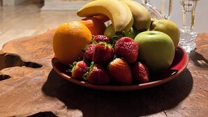 A fruit bowl on a wooden table with a stone wall and a bed in the background.