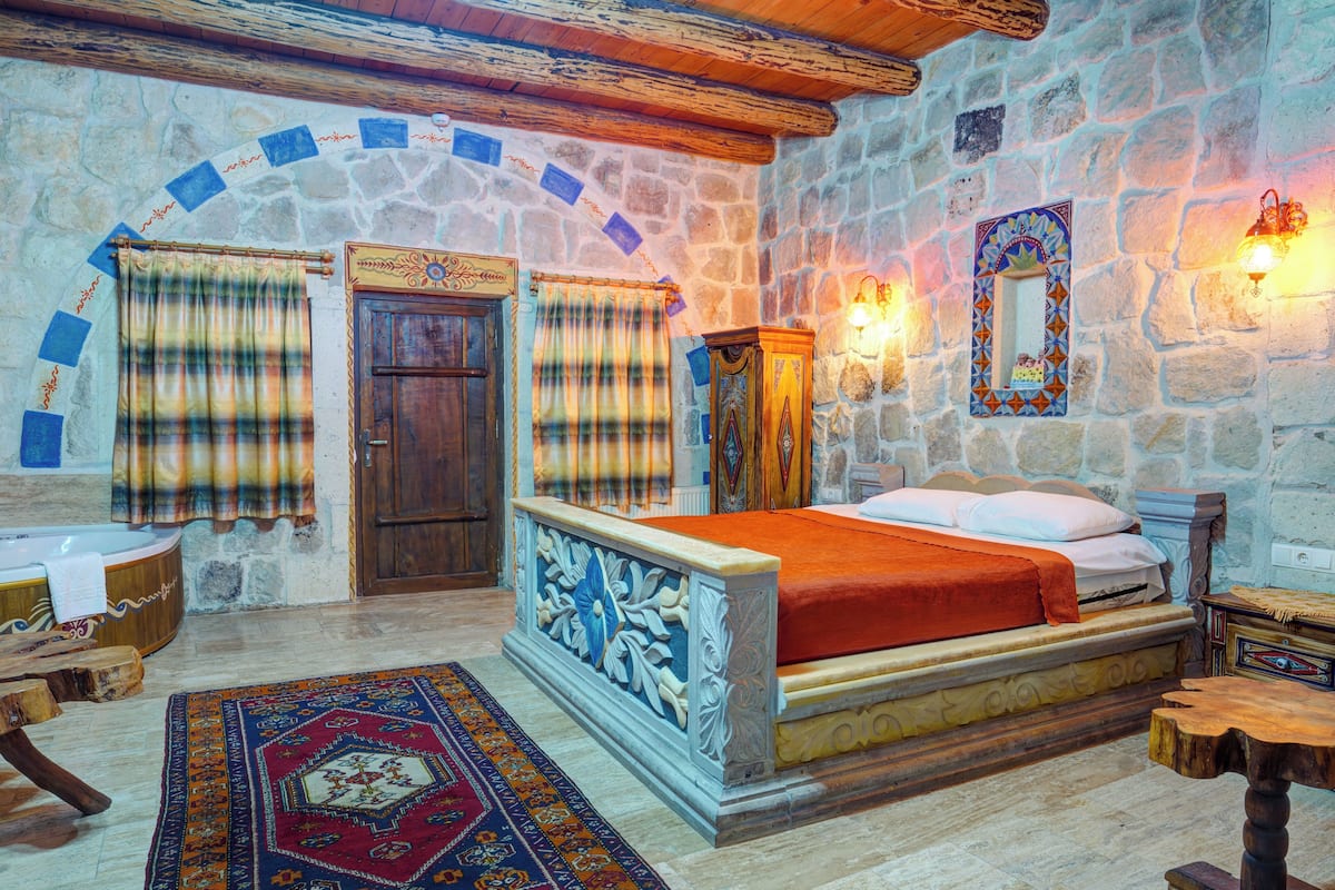A stone-walled bedroom with a bed, curtains, a wooden wardrobe, a mirror, and a small table.