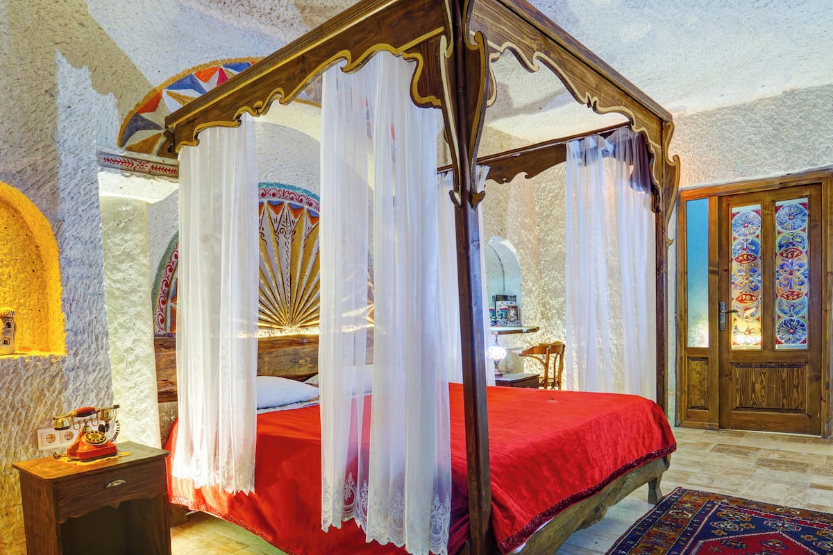 A four-poster bed with a red bedspread and white drapes, a wooden nightstand with a telephone, and a patterned rug on the floor.