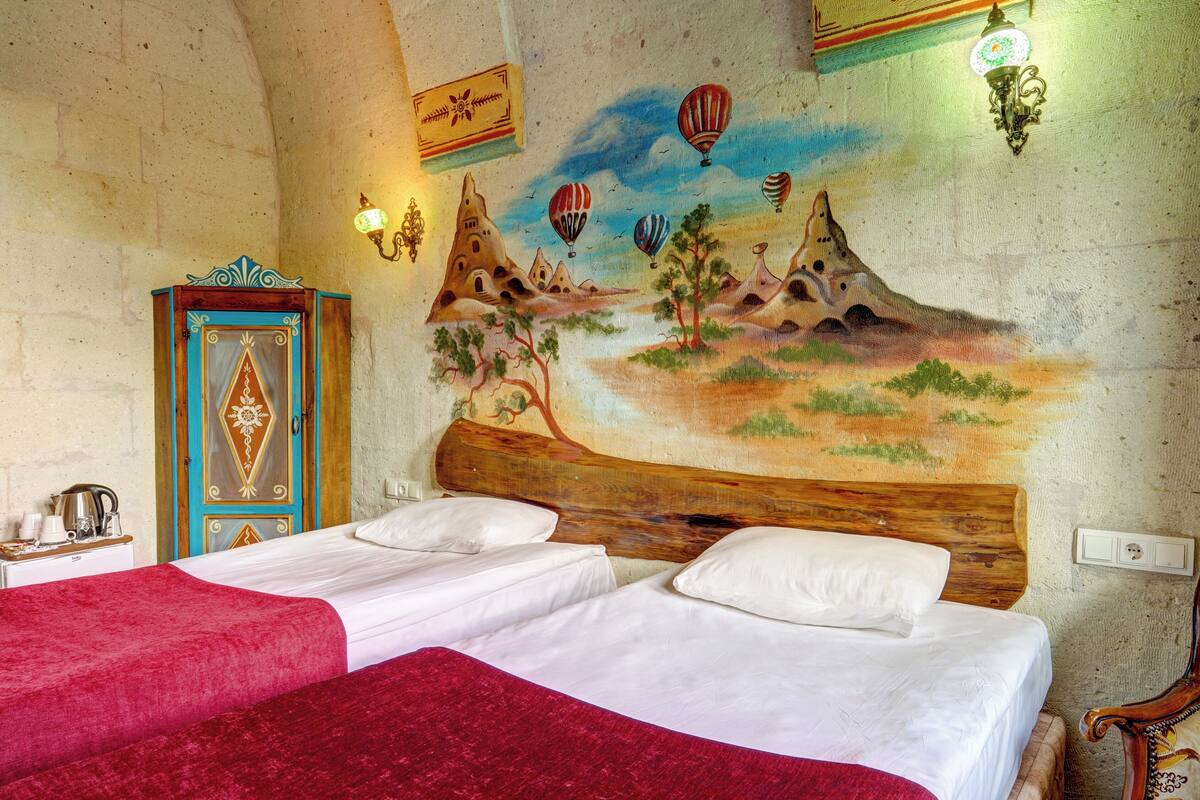 A room with a bed, a wooden headboard, a red bedspread, a wall with a mural, and a blue cabinet.