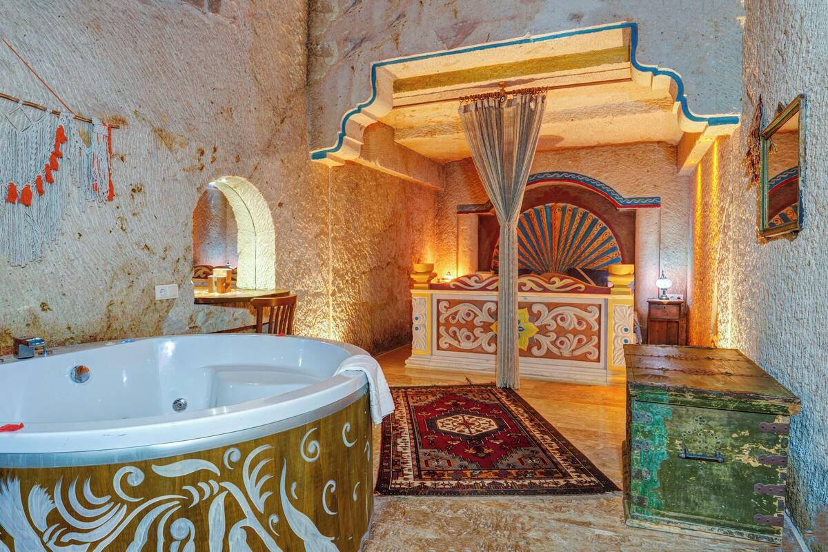 A room with a large bathtub, a bed with ornate headboard, and a small table with a lamp.