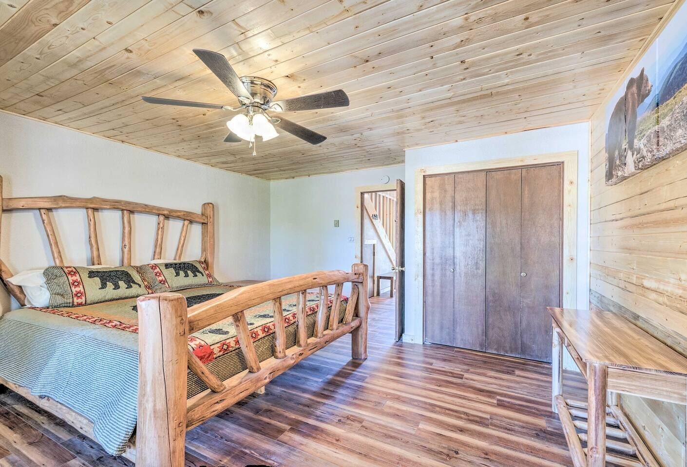 A wooden-floored bedroom with a bed, a wooden headboard, a ceiling fan, and a door.