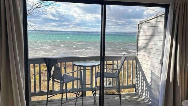 Quarto com uma grande porta de vidro deslizante que dá para uma varanda com vista para o oceano. Há uma cama com colcha acolchoada e uma pequena mesa redonda com cadeiras na varanda.