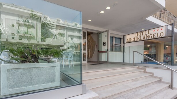 Ein moderner Hotelaufgang mit Glasfassade, einer Treppe und einem Schild mit der Aufschrift 'HOTEL MICHELANGELO'.