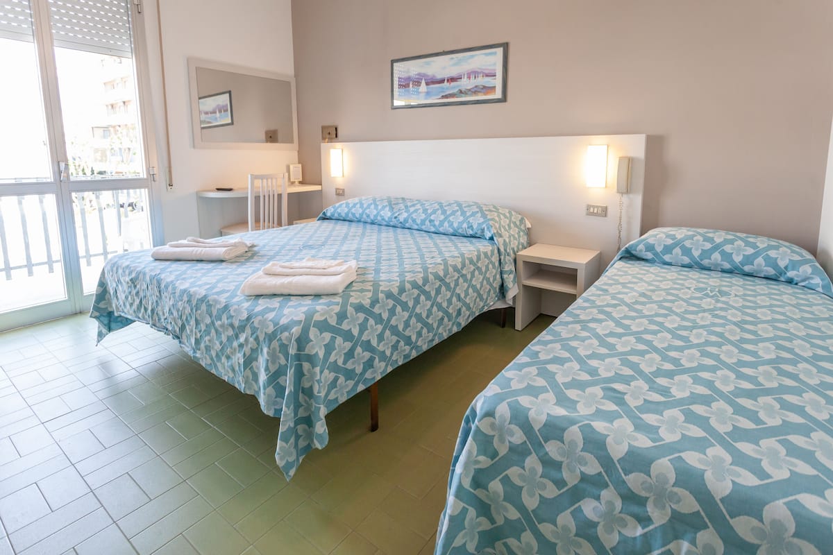 Ein Hotelzimmer mit zwei Betten, jedes mit einer blauen, floralen Bettdecke und weißen Kissen. Es gibt zwei Nachttische mit Lampen, einen kleinen Tisch und einen Sessel. Ein Gemälde hängt an der Wand, und ein Fenster bietet Blick auf Gebäude.