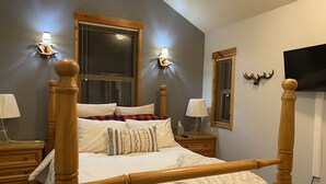 Un lit à baldaquin avec une structure en bois, une couverture blanche moelleuse et une couverture à carreaux. Il y a deux tables de chevet avec des lampes et un téléviseur fixé au mur.