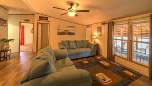 Una sala de estar con un sofá grande, un ventilador de techo, un cuadro en la pared y vistas por la ventana.