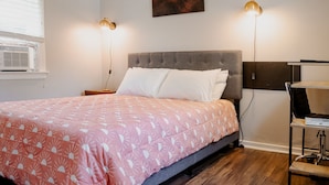 A neatly made bed with a pink and white patterned bedspread, two bedside lamps, a gray upholstered headboard, and a painting of a sunset on the wall.