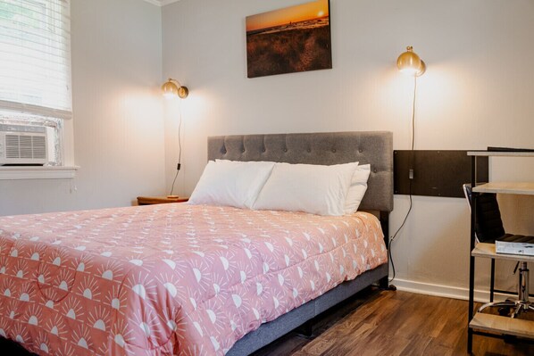 A neatly made bed with a pink and white patterned bedspread, two bedside lamps, a gray upholstered headboard, and a painting of a sunset on the wall. - Relaxed 1 BR Near Lake with Fast WiFi, Fire Pit + (Coloma)