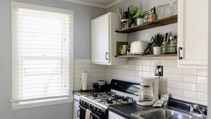 A kitchen with a gas stove, a coffee maker, and a sink. There is a window with blinds and a shelf with kitchen utensils and plants.