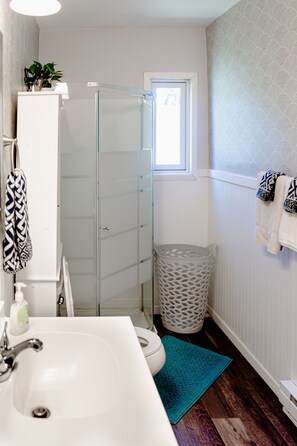 A modern bathroom with a glass shower enclosure, a white sink, a blue rug, and a patterned trash bin. - Relaxed 1 BR Near Lake with Fast WiFi, Fire Pit + (Coloma)