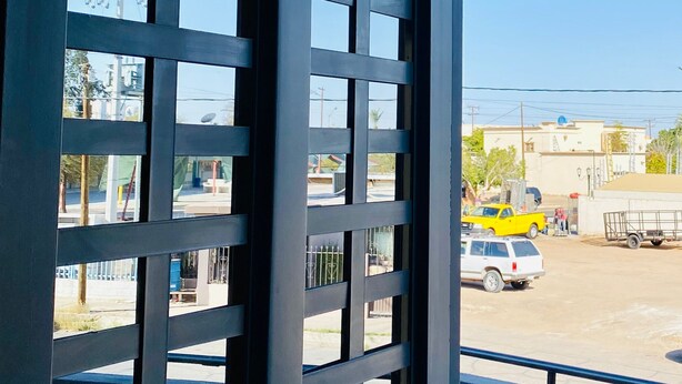 A view from a balcony with a metal grid door, a clear blue sky, and a parking lot with vehicles. - Hotel Santa Cruz Residencial (San Luis Rio Colorado)