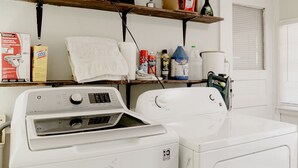 Un espace buanderie comprenant un lave-linge et un sèche-linge à chargement frontal, une étagère avec des produits de nettoyage et une fenêtre avec des stores.