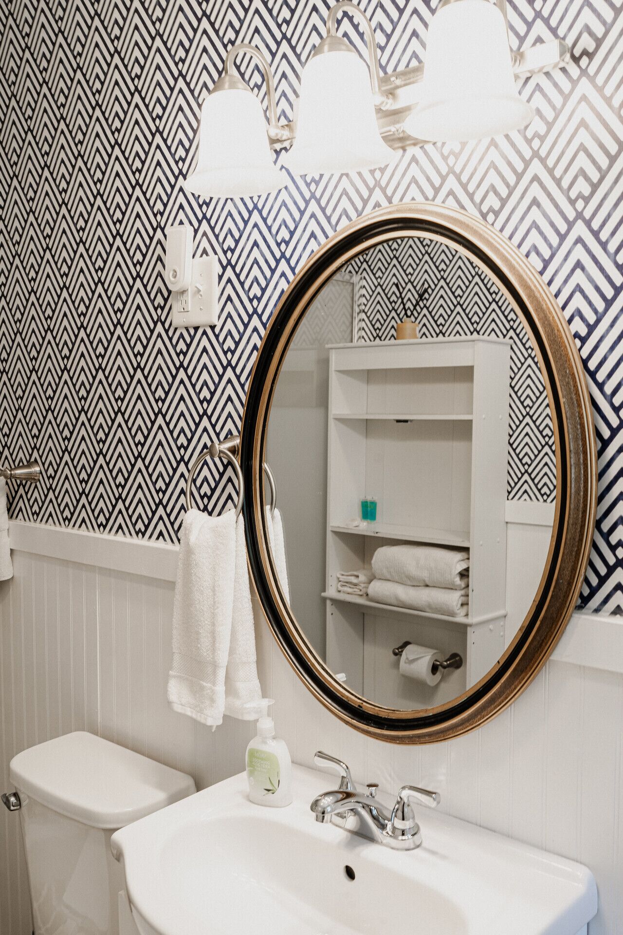 Et badeværelse med en hvid vask, toilet, et spejl med gylden ramme og tapet.