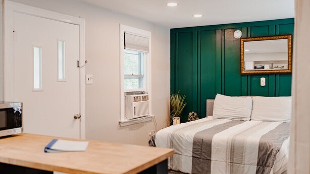 Une chambre à coucher comprenant un lit, un bureau, une fenêtre avec des stores, un mur d’accent de couleur verte et un miroir.