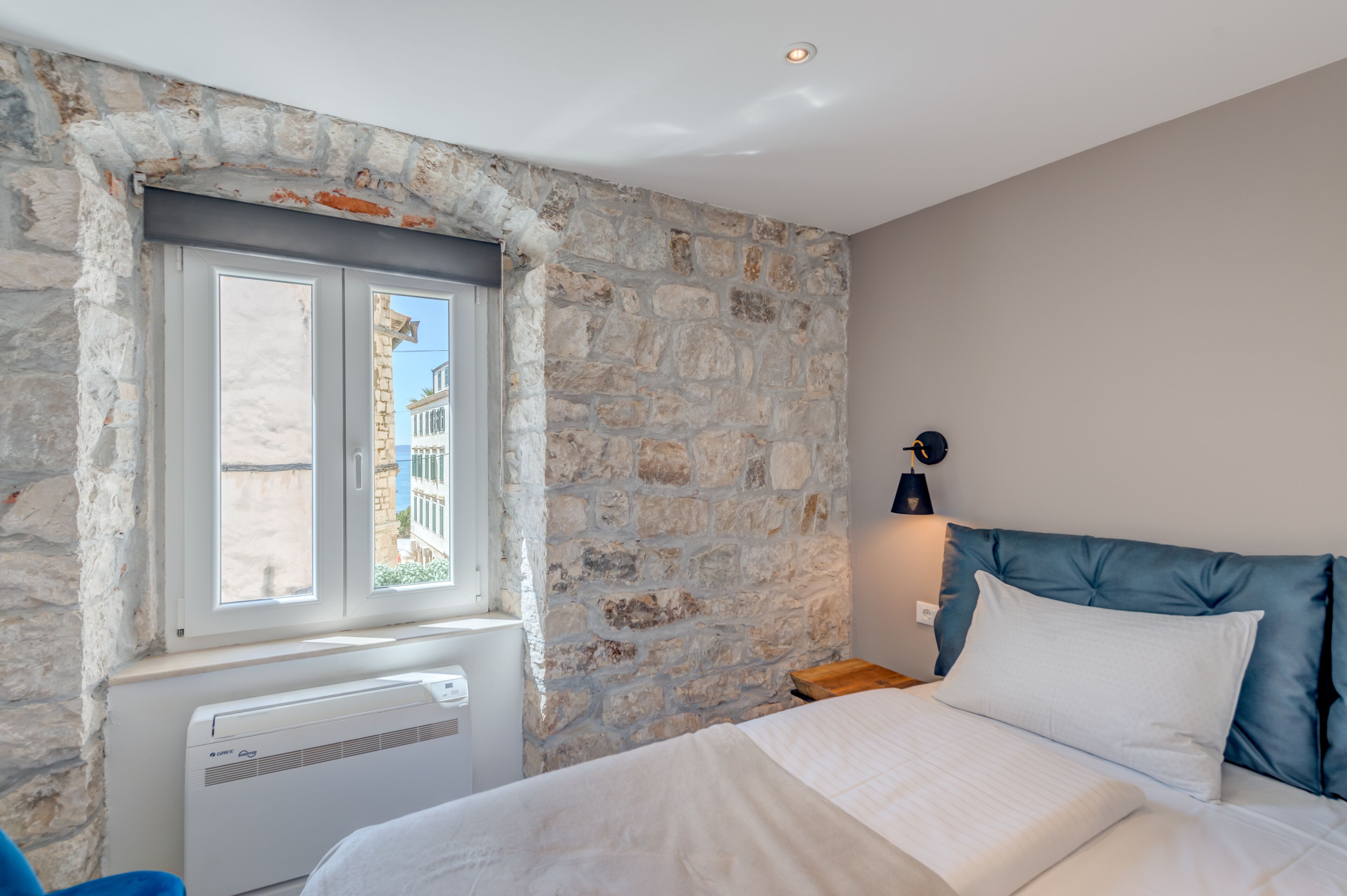 A bedroom with a stone wall, a window, a bed with white bedding, and a nightstand with a lamp.
