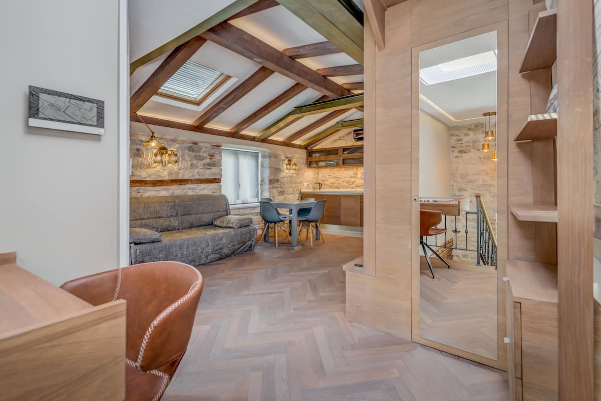 A cozy loft-style room with exposed wooden beams, a stone wall, and a herringbone-patterned floor.