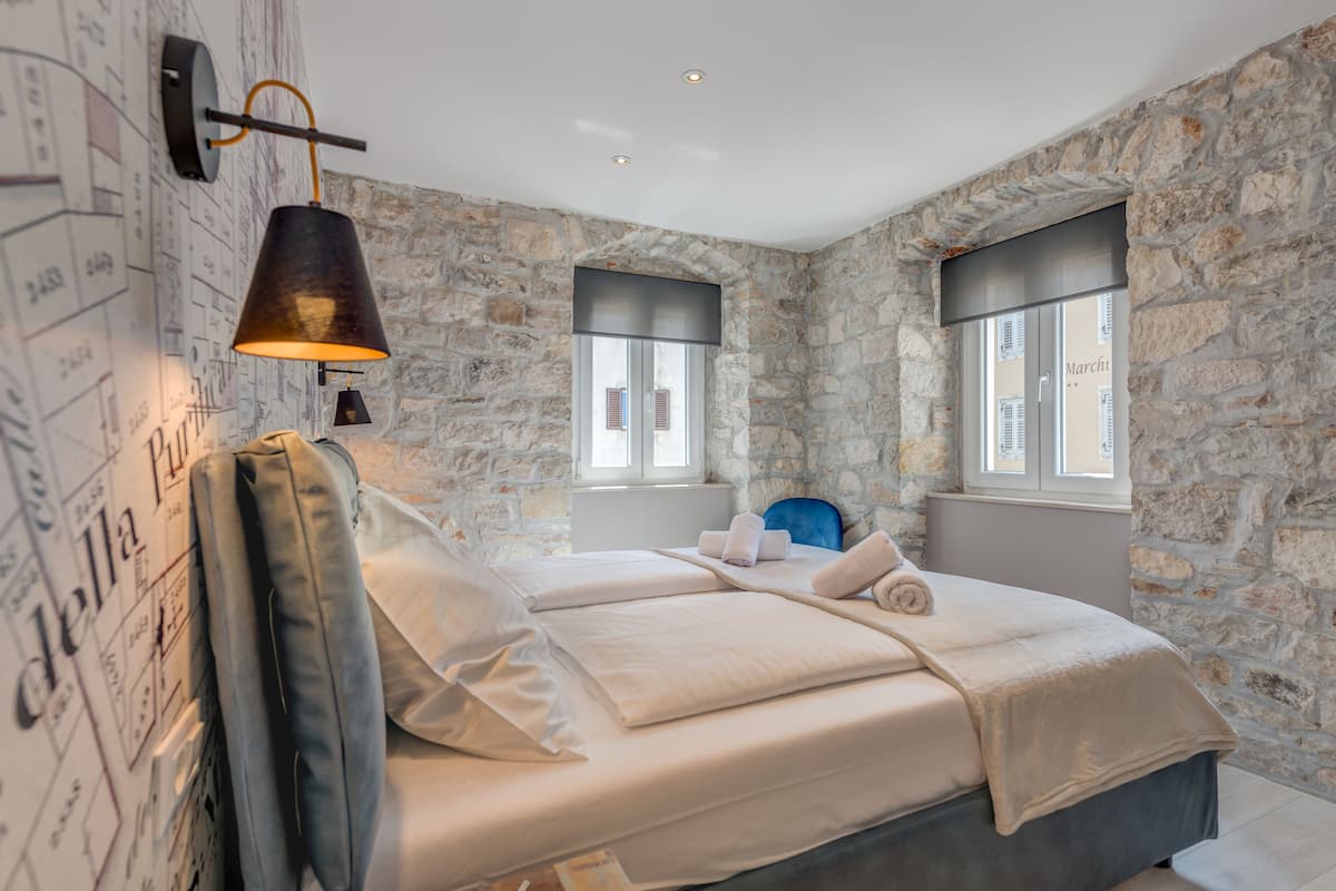 A bedroom with a stone wall, a bed with white linens, a hanging lamp, and two windows.