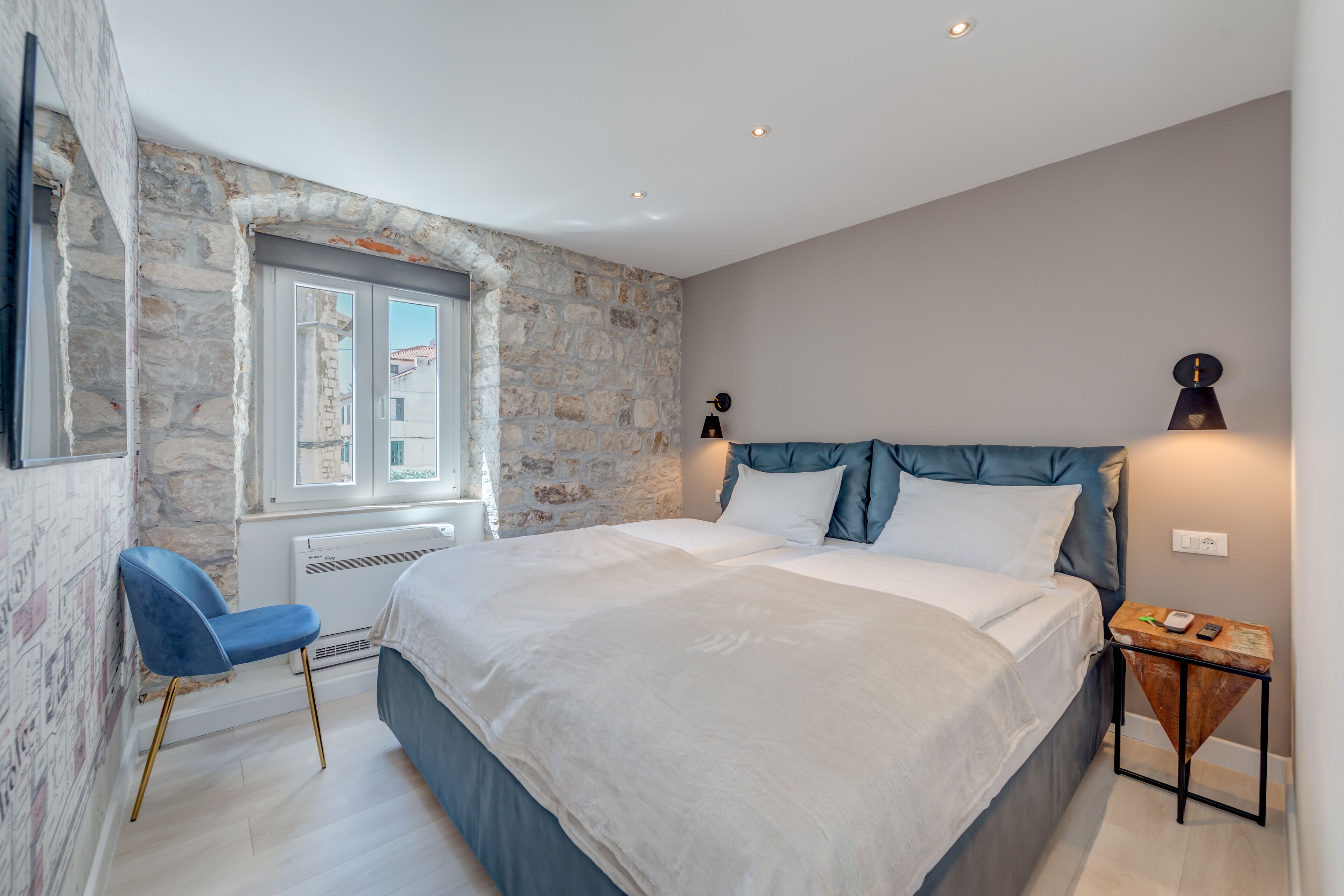 Quarto com parede de pedra, cama com roupa de cama branca, uma cadeira azul e mesa de cabeceira com abajur.