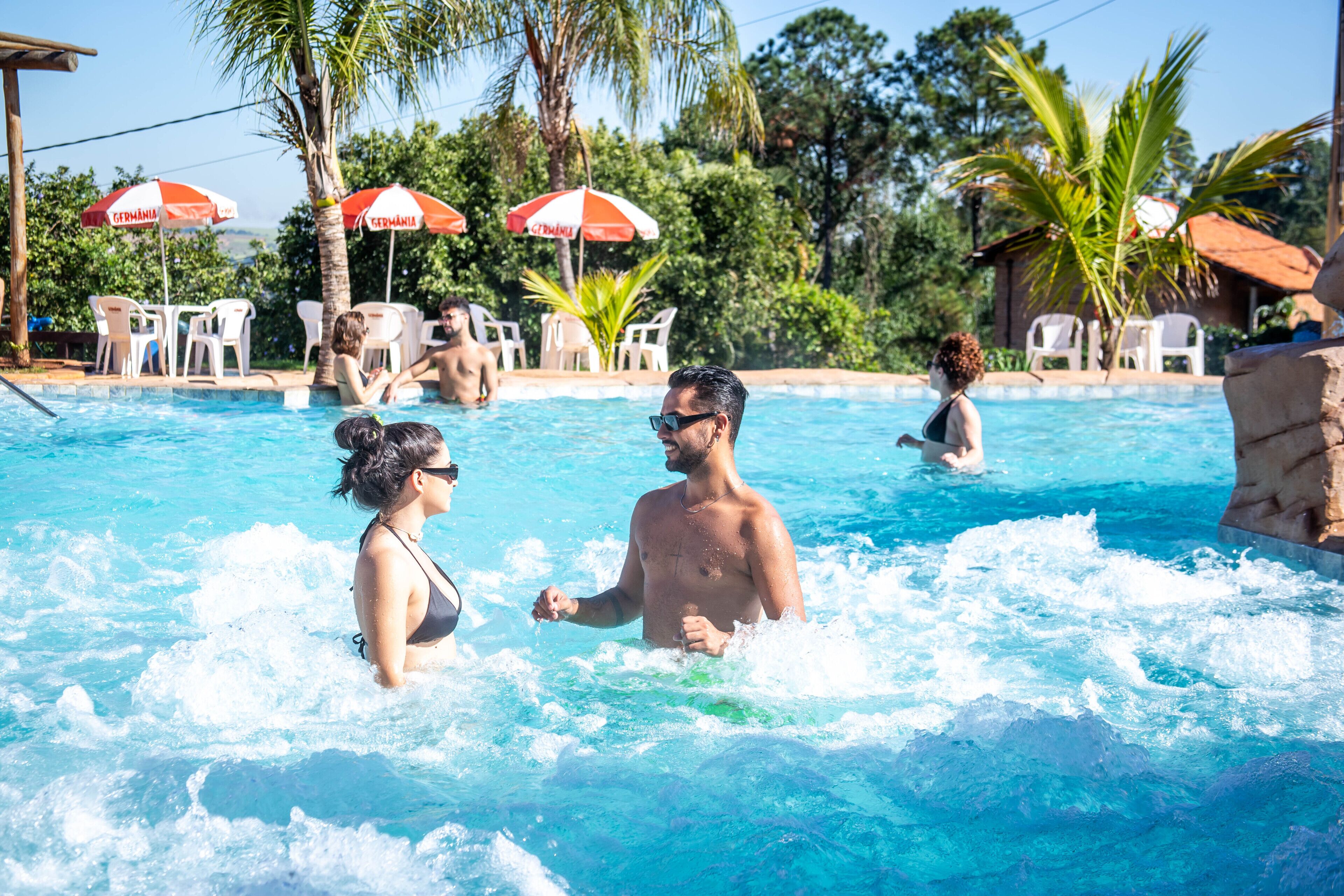 A swimming pool with people in the water, surrounded by palm trees and umbrellas.