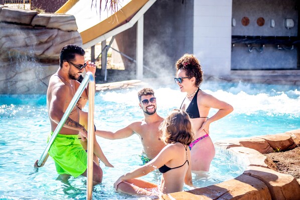 A group of people in a swimming pool with a slide and rocky edges.