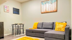 A living room with a gray sectional sofa, a yellow throw rug, and a painting on the wall. - Newly Built and Cheerful 1-Bedroom Suite (Calgary)