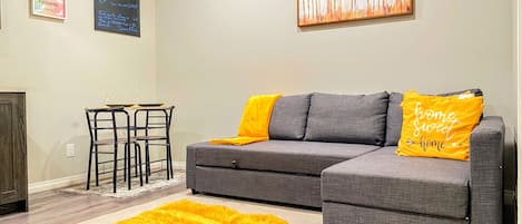 Un salon avec un canapé d’angle gris, un tapis jaune, une table à manger avec des chaises et un tableau accroché au mur.