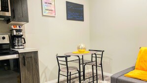 A compact kitchen with a coffee maker, a small table with chairs, and a sofa with a yellow throw. - Newly Built and Cheerful 1-Bedroom Suite (Calgary)