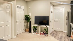 A compact living space with a flat-screen TV on a white stand, a refrigerator, and a door with a 'Welcome' sign. - Newly Built and Cheerful 1-Bedroom Suite (Calgary)