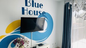 Een kamer met een wandgemonteerde flatscreen-tv, een witte console tafel met een bloemenvaas en een groot raam met blauwe gordijnen.